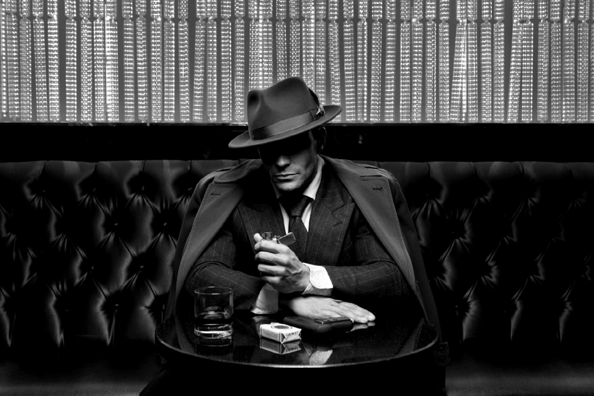 Mann im Anzug und Hut sitzt in Schwarzweiß an einem Tisch mit Glas, Zigarettenpackung und Feuerzeug